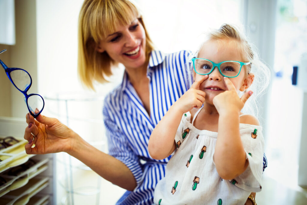 Happy mother with her cute daughter choosing glasses in optics store